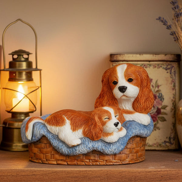 A decorative figurine of two Cavalier King Charles Spaniel dogs resting in a blue blanket on a woven basket base