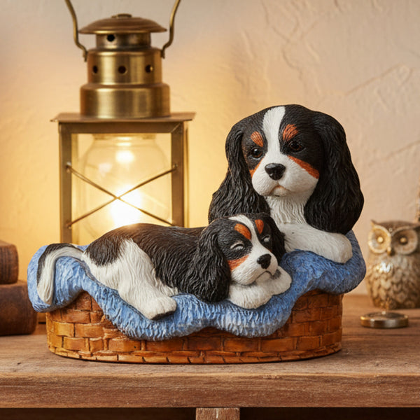 A decorative figurine of two black-and-white Cavalier King Charles Spaniel dogs resting on a blue cushion inside a brown basket, 