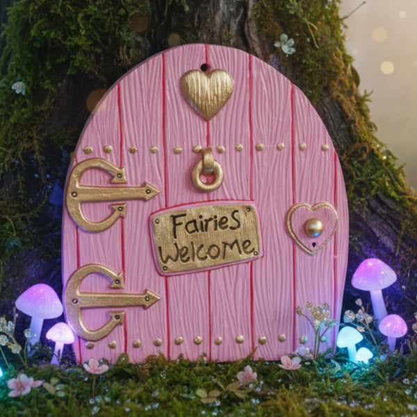 Pink, arched fairy door with a wood-grain texture. The door is decorated with gold hinges, a heart-shaped symbol, a ring knocker, and a heart-shaped keyhole. A gold plaque in the centre reads, "Fairies Welcome."