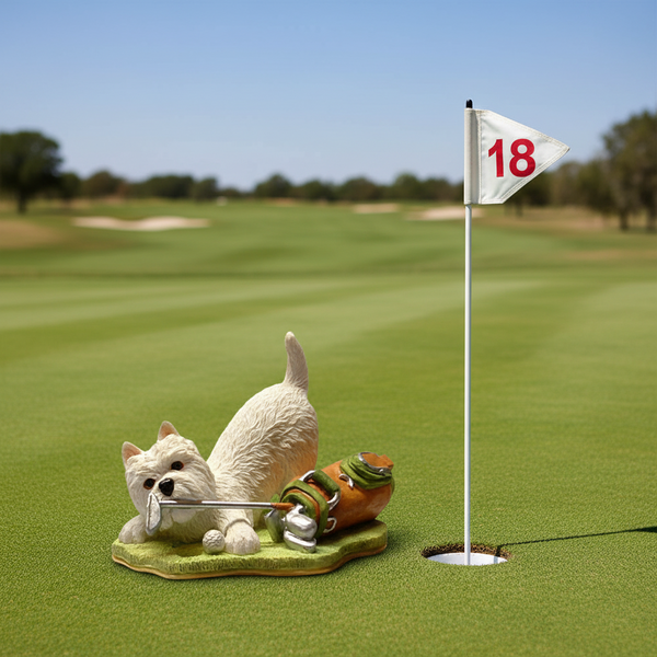 A small figurine of a westie lying on a green base, holding a silver-colored golf club with a golf ball, next to a small  golf bag